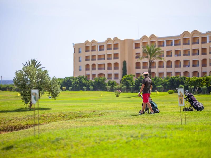 Dos personas jugando al golf en un campo verde frente a un gran edificio hotelero.