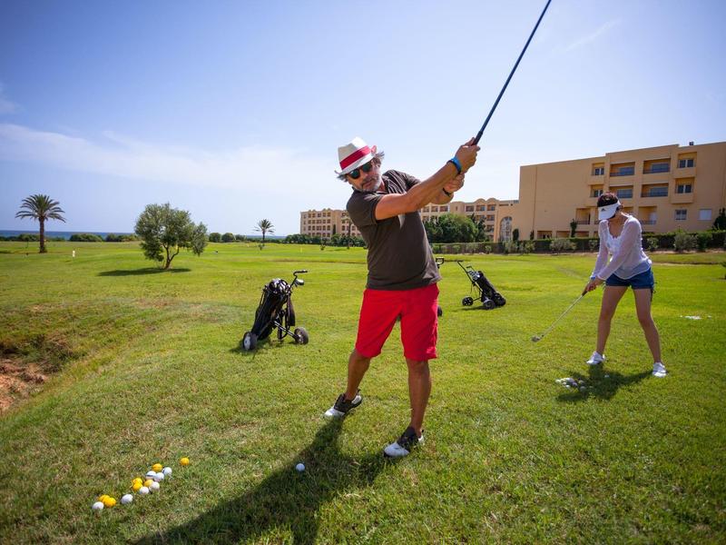 Dos personas jugando golf en un campo con césped, edificio y cielo despejado al fondo.