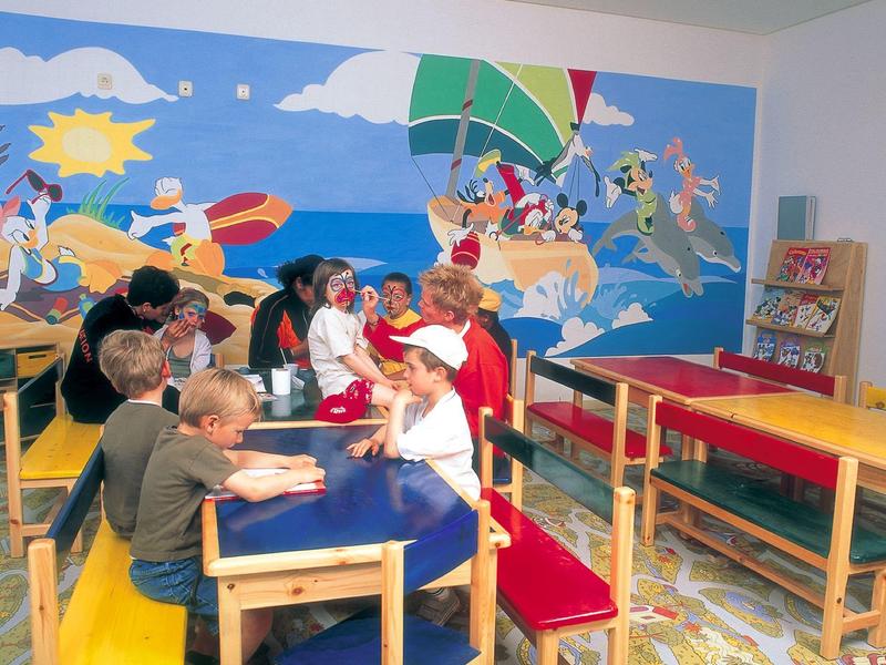 Niños sentados en mesas coloridas en una sala con una gran pintura mural del mar.