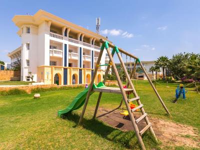 Children's playground with slide and climbing frame near a multi-story hotel building in sunny weather.