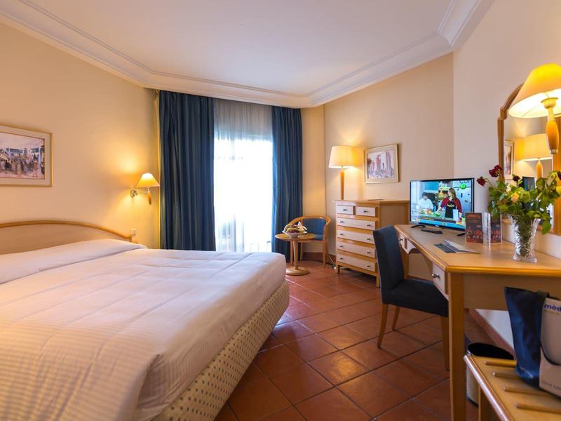 Bright hotel room with double bed, desk, TV, and window with curtains.