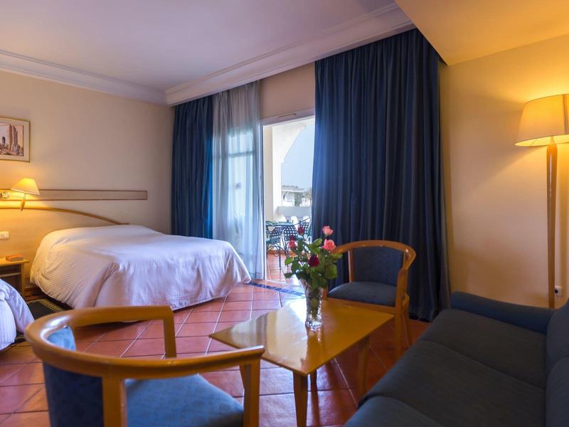 Cozy hotel room with double bed, sofa, armchair, and a table with flowers.