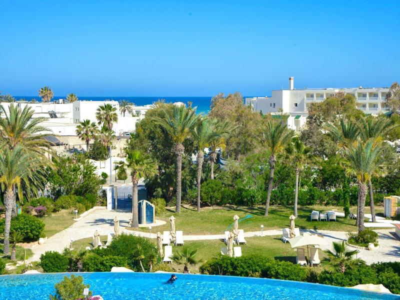 View of a hotel garden with palm trees, white buildings, and the sea in the background.