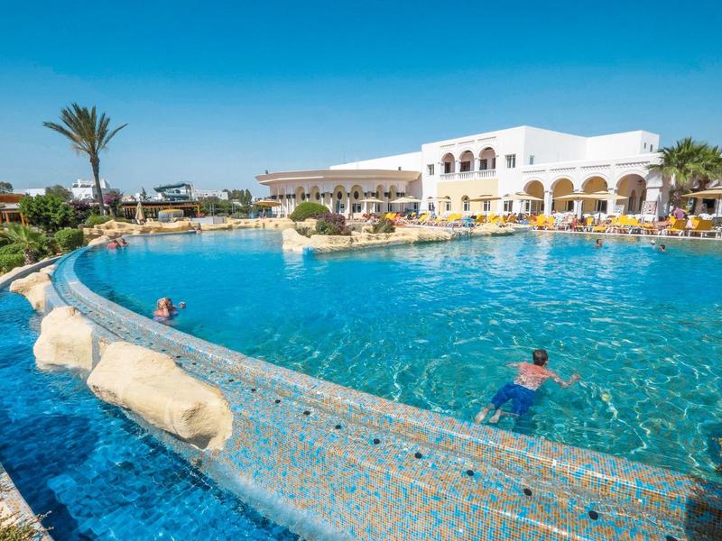 Large pool in front of a white hotel building with sun umbrellas and palm trees under a blue sky.