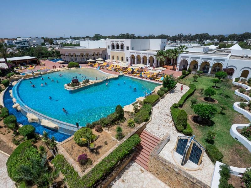 Large hotel pool with people, surrounded by manicured gardens and white architecture under a blue sky.