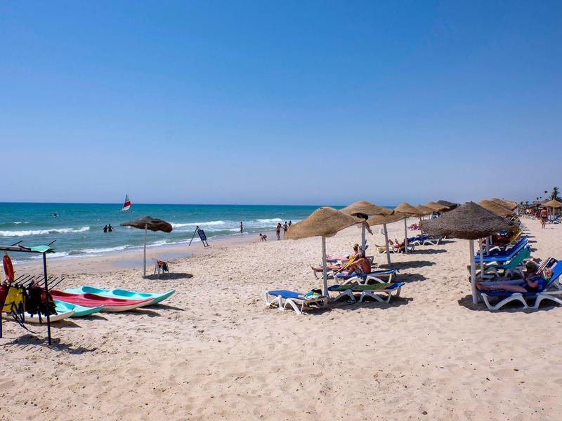 Beach with lounge chairs, straw umbrellas, surfboards, clear sky and sea.