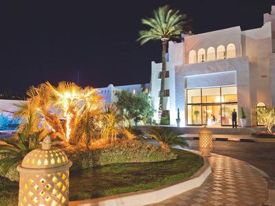 Beleuchtetes Hotelgebäude mit Palmen und Gartenanlage bei Nacht.