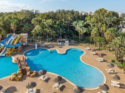 Großer Hotelpool mit Wasserrutschen und Sonnenliegen, umgeben von Palmen und Gebäuden.