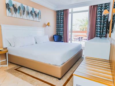 Hell eingerichtetes Hotelzimmer mit großem Bett, Sitzbereich und Balkon mit Blick ins Grüne.