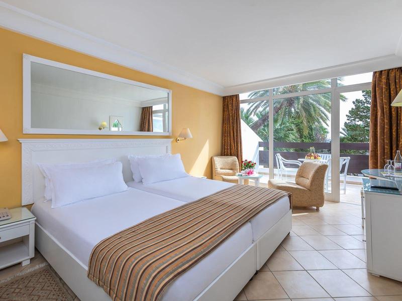 Helles Hotelzimmer mit Doppelbett, Spiegel, Sesseln und Zugang zur Terrasse mit Palmenblick.