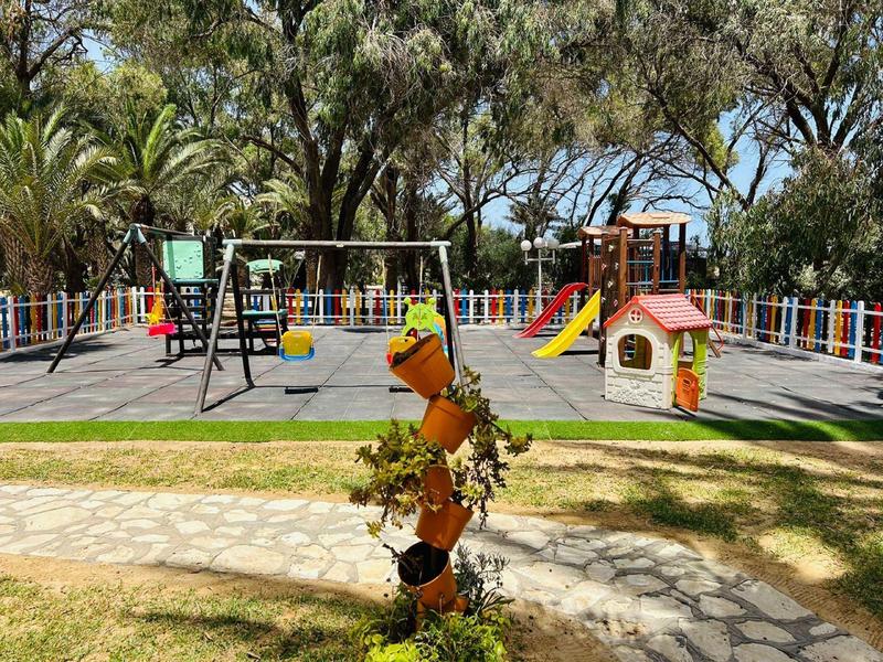 Spielplatz mit Schaukeln, Rutschen und einem kleinen Spielhaus, umgeben von Bäumen.