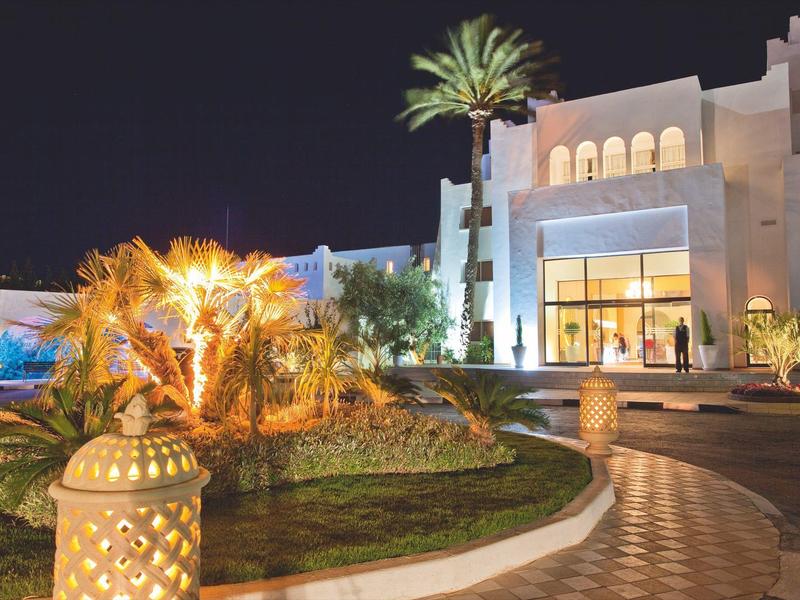 Beleuchtetes Hotelgebäude mit Palmen und Gartenanlage bei Nacht.