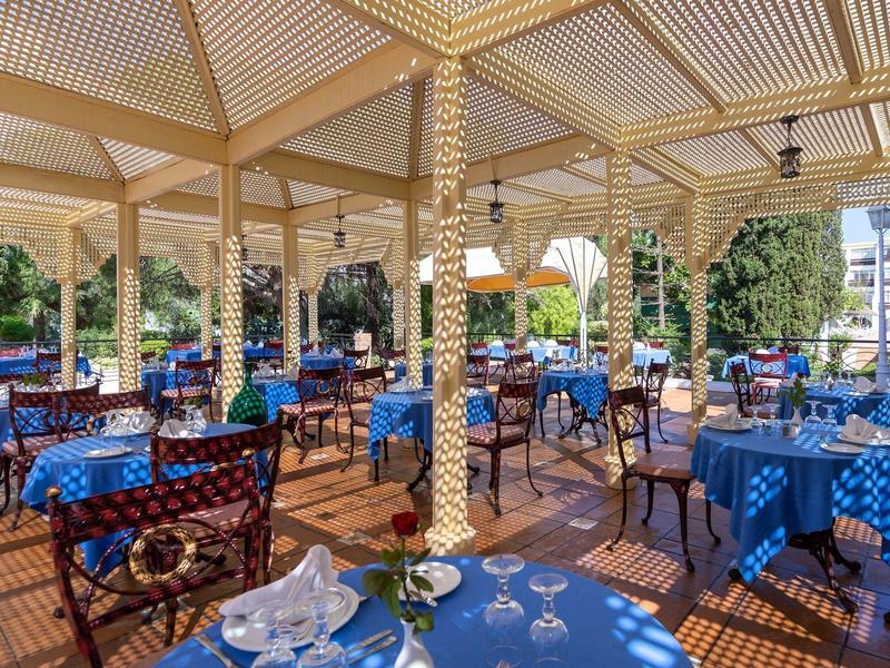 Offener Restaurantbereich mit blauen Tischtüchern und hölzernen Stühlen unter einem Holzpergola-Dach.
