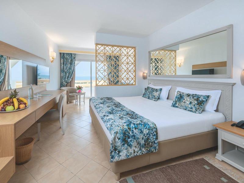 Helles Hotelzimmer mit Doppelbett, Schreibtisch, Stühlen und großem Fenster mit Meerblick.