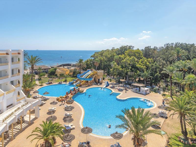 Großer Hotelpool mit Liegestühlen, Palmen und Meerblick bei klarem Himmel.