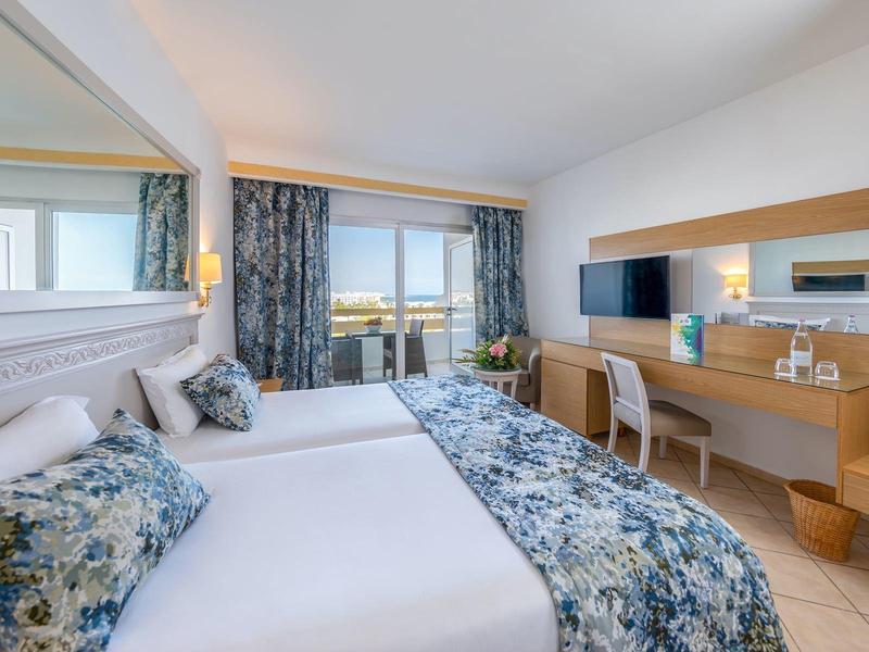 Helles Hotelzimmer mit Doppelbett, Schreibtisch und Balkon mit Meerblick.