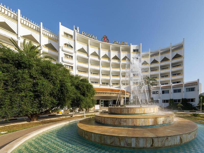 Hotel mit mehrstöckigem Gebäude, Brunnen und klar blauem Himmel im Urlaubsumfeld.