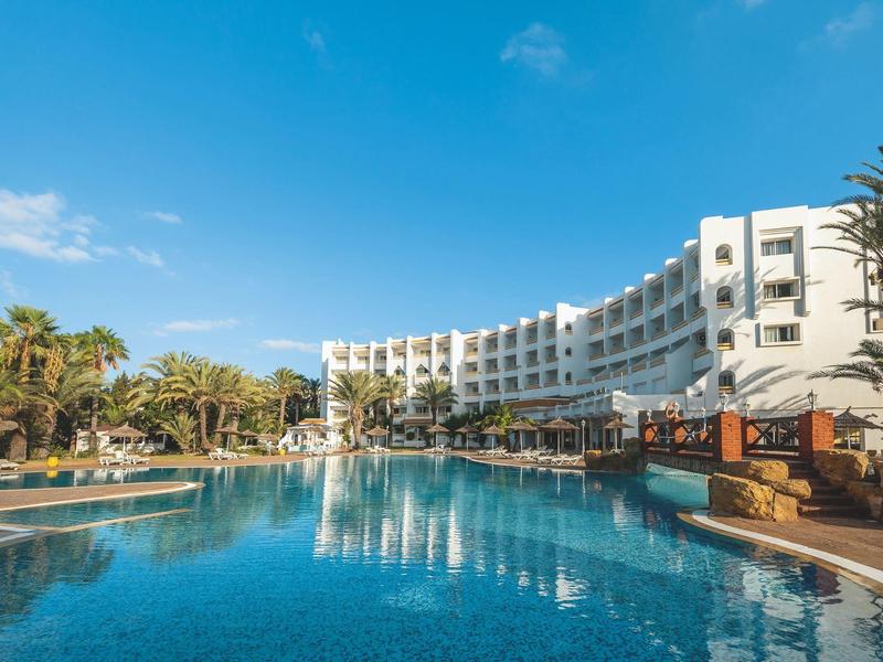 Großer Hotelpool mit klarem Wasser vor einem weißen mehrstöckigen Hotel und Palmen bei blauem Himmel.