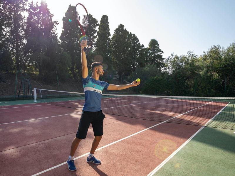 Mann spielt Tennis auf einem sonnigen Außenplatz mit Bäumen im Hintergrund.