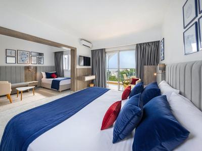 Jasny pokój hotelowy z dużym łóżkiem, niebieskimi i czerwonymi poduszkami oraz widokiem na taras.