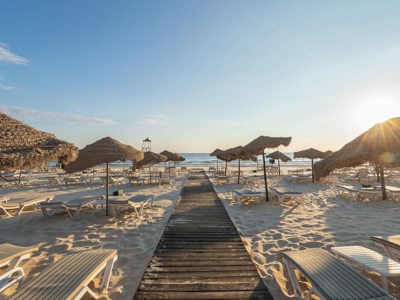 Piaszczysta plaża z leżakami i parasolami o zachodzie słońca, drewniana ścieżka prowadząca do morza