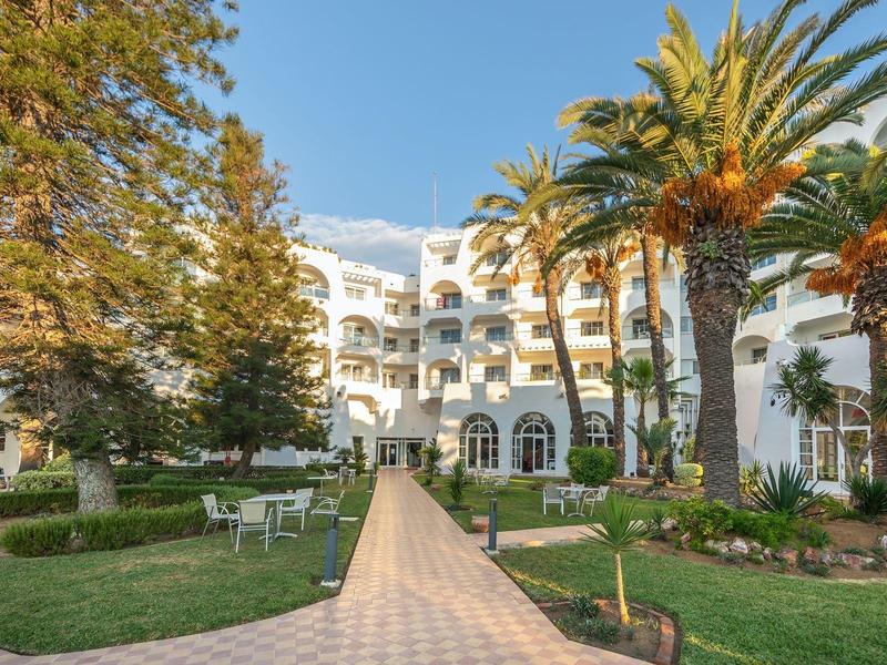 Biały budynek hotelowy z balkonami, otoczony palmami i zielonymi ogrodami pod niebieskim niebem.