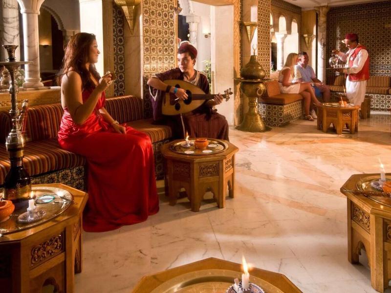 Útulný salonek s lidmi užívajícími si nápoje a kytarovou hudbu v elegantním, teple osvětleném prostoru.