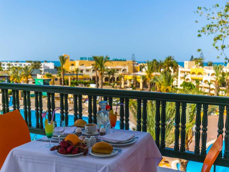 Stůl na balkóně s jídlem, výhled na palmy a žluté budovy pod jasnou modrou oblohou.