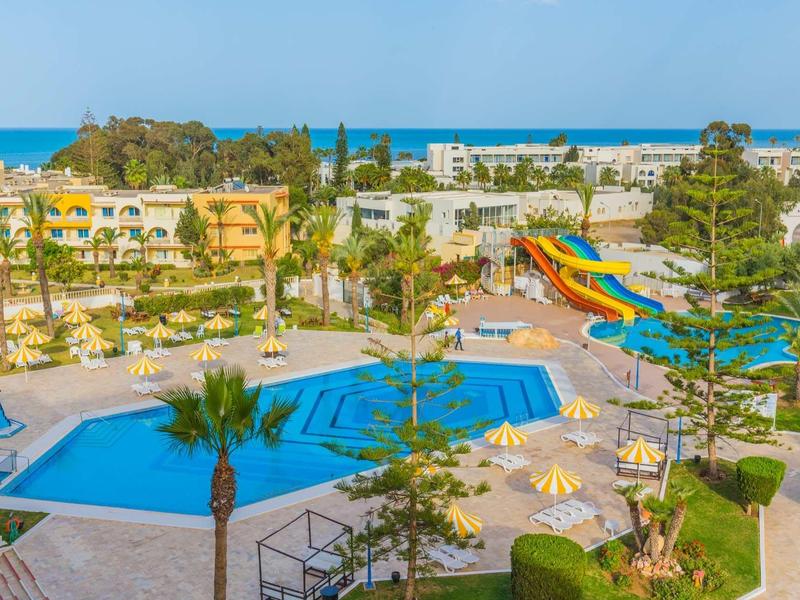 Slunečné letovisko s bazény, skluzavkami, palmami a mořem v dálce.