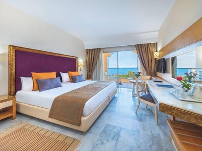 Helles Hotelzimmer mit großem Bett, lila Kopfteil, Schreibtisch, Meerblick und Balkon.