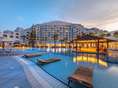 Großer Hotelpool mit Liegen und Palmen bei blauem Himmel im abendlichen Licht.