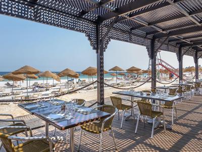 Terrazza coperta sulla spiaggia con tavoli e sedie, vista su lettini e ombrelloni vicino al mare.