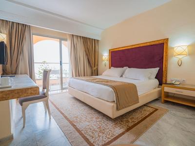 Camera d'hotel luminosa con letto matrimoniale, comodino, scrivania e balcone con vista sul mare.