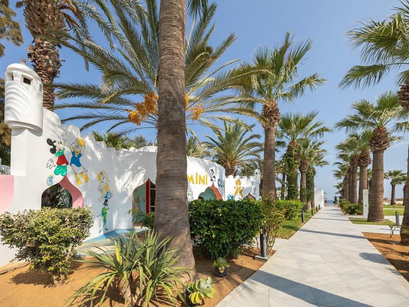 Sentiero fiancheggiato da palme con edificio bianco decorato con mosaici colorati.