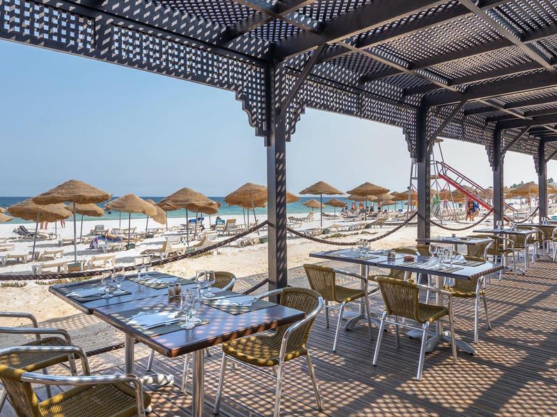 Terrazza coperta sulla spiaggia con tavoli e sedie, vista su lettini e ombrelloni vicino al mare.