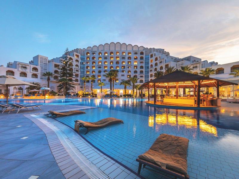 Grande piscina dell'hotel con lettini e bar piscina al tramonto.
