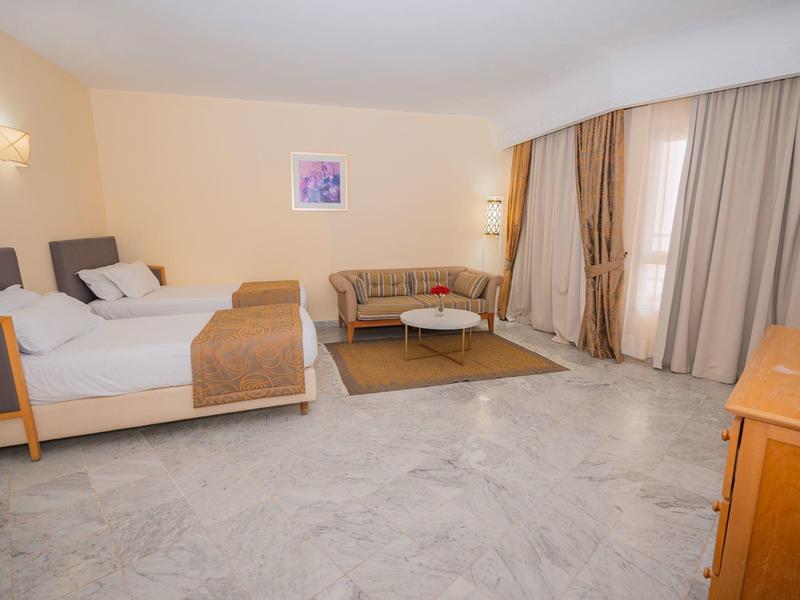 Camera d'hotel luminosa con due letti, divano, tappeto e grandi finestre con tende.