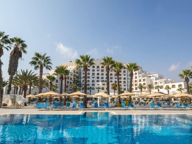 Grande hotel a più piani accanto a una piscina con palme sotto un cielo azzurro