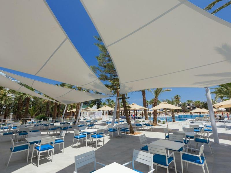 Area esterna di un hotel con tavoli, sedie e grandi tende da sole sotto un cielo sereno.