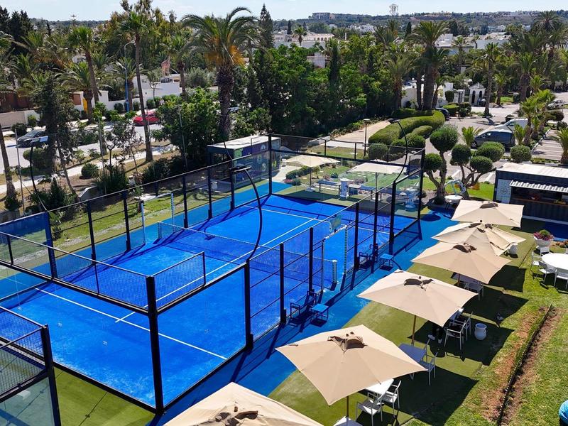 Campi da paddle tennis blu con posti a sedere e ombrelloni in un ambiente vacanziero.