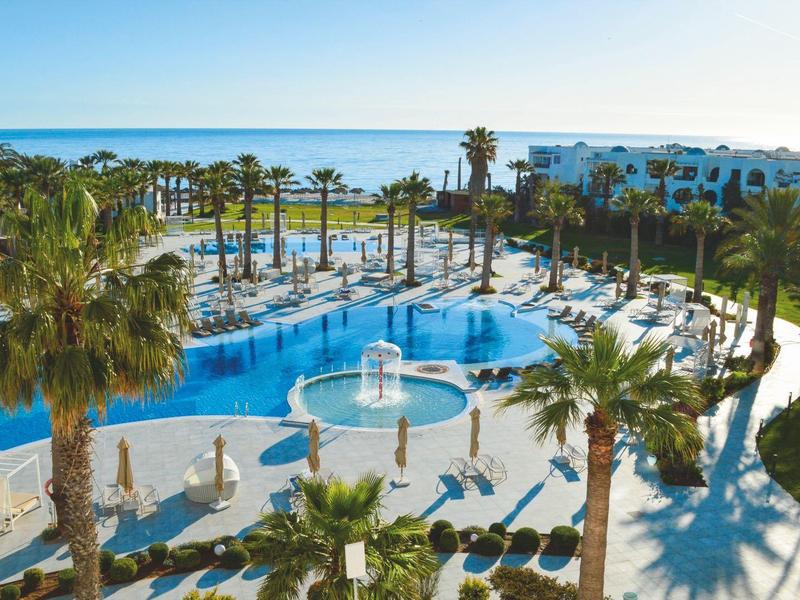 Piscina esterna con palme, lettini e vista sul mare in un hotel.