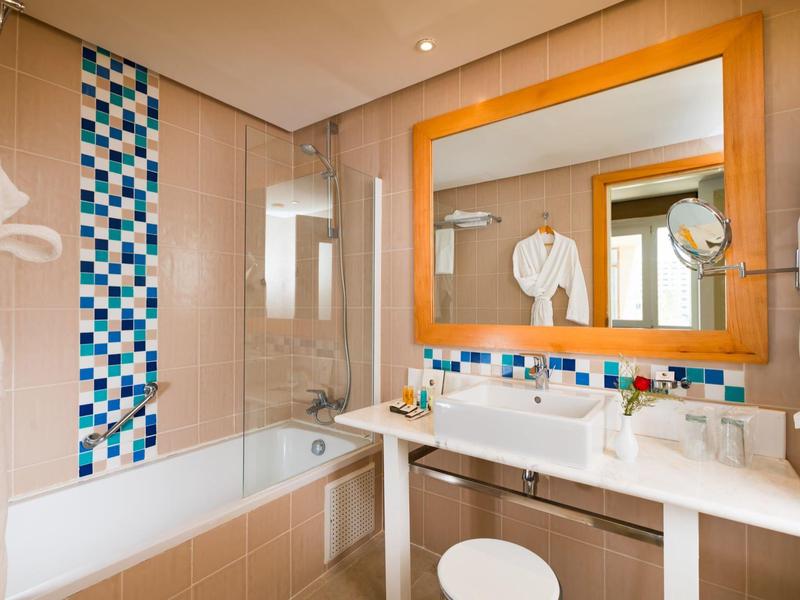 Jasna łazienka z białą umywalką, dużym lustrem i wanną z zasłoną prysznicową.