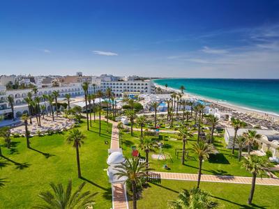 Pohled na pobřežní hotelový komplex se zelenými zahradami, cesty lemované palmami a pláží u moře.