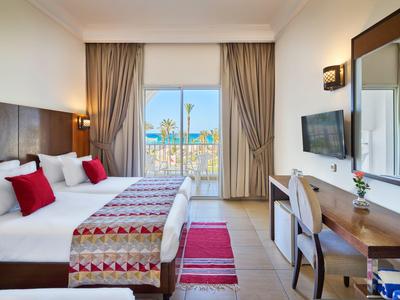 Světý hotelový pokoj s manželskou postelí, dřevěným nábytkem a balkonem s výhledem na palmy.