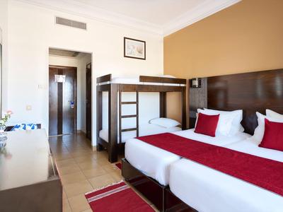 Hotelový pokoj s manželskou postelí, palandou, pracovním stolem a teplými odstíny.