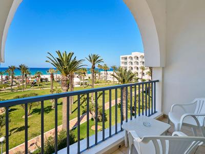 Balkon s bílými židlemi s výhledem do zahrady, na palmy a moře pod modrou oblohou.