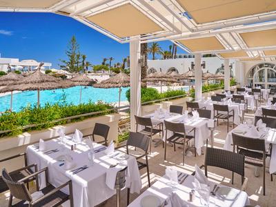 Venkovní restaurace s prostřenými stoly vedle bazénu pod velkou sluneční clonou.