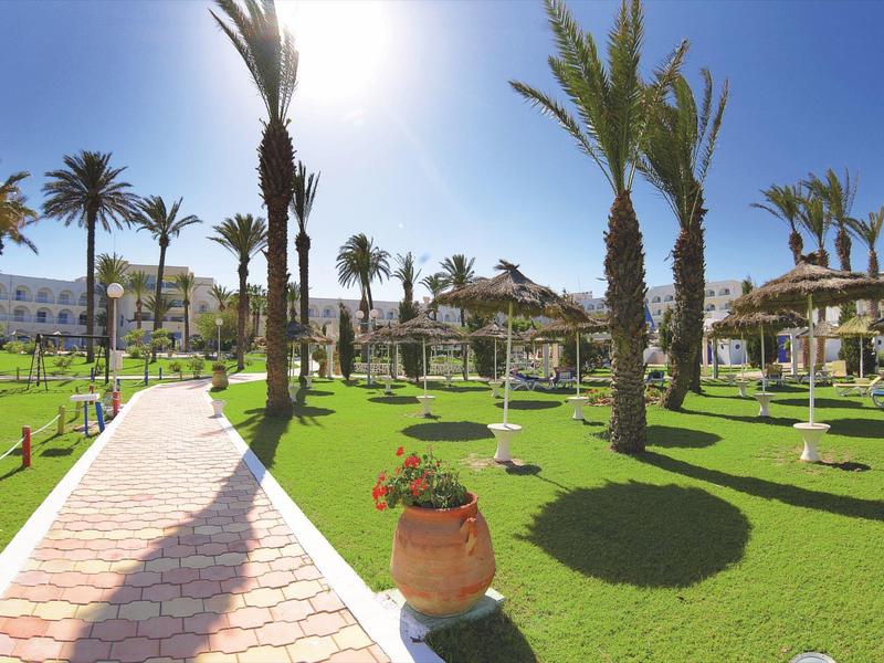 Grand domaine hôtelier avec palmiers, pelouses vertes et chemin pavé sous un soleil éclatant.