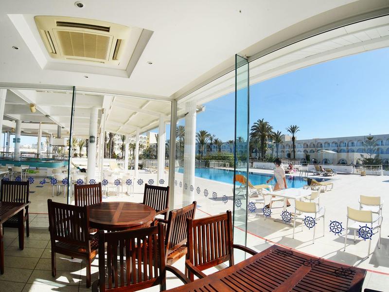 Espace intérieur d'hôtel avec tables et chaises donnant sur une piscine et des parasols.