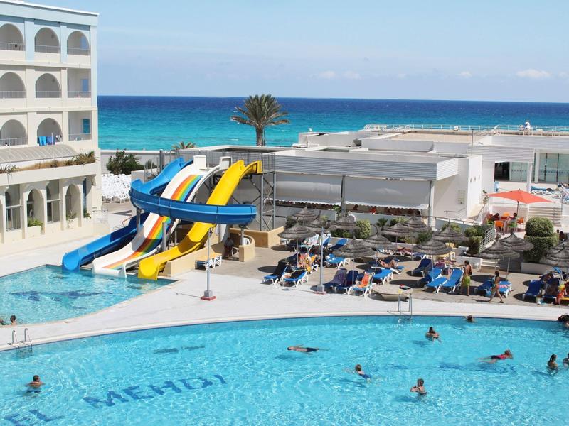 Grand piscine d'hôtel avec toboggans aquatiques et vue sur la mer en arrière-plan.